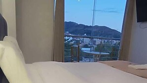 A hotel room with a bed, a bedside lamp, a view of a cityscape and mountains through a large window.