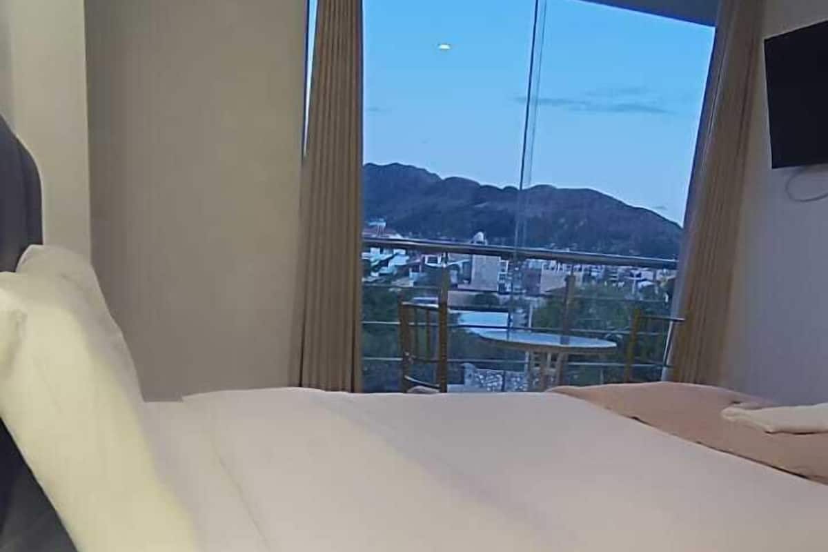 A hotel room with a bed, a bedside lamp, a view of a cityscape and mountains through a large window.