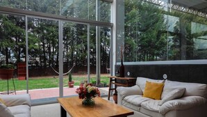 Een moderne woonkamer met een groot raam, een houten salontafel, een witte bank met een geel kussen en een kleine tafel met een bloemenvaas.