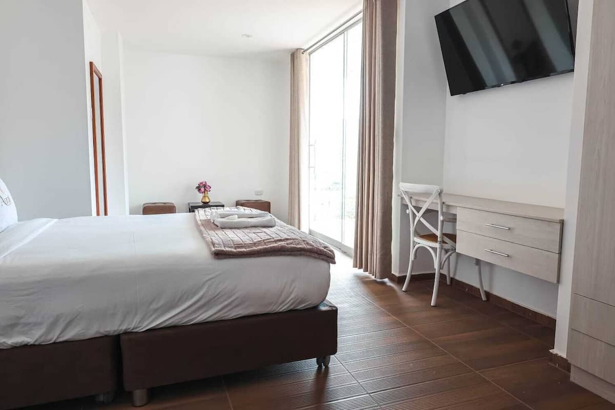 A hotel room with a large bed, a desk with drawers, a chair, a TV mounted on the wall, and a window with curtains.