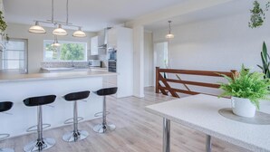 A modern kitchen with a central island, bar stools, pendant lighting, and a wooden dining table with a potted plant.