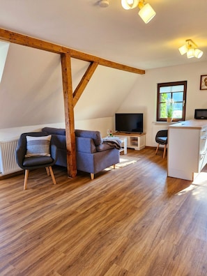 En moderne stue med en træbjælke i loftet, en grå sofa, et lille bord med et fjernsyn og et vindue med en plante.