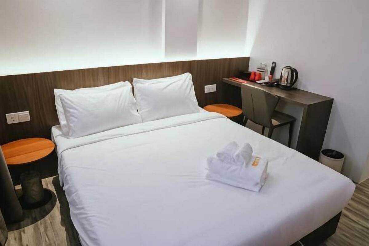 A hotel room with a bed, a desk, a chair, and a small table with a kettle.