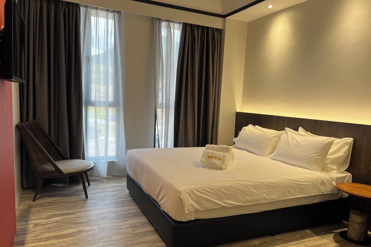 A hotel room with a large bed, a chair, a bedside table, and a view of the outdoors through the window.