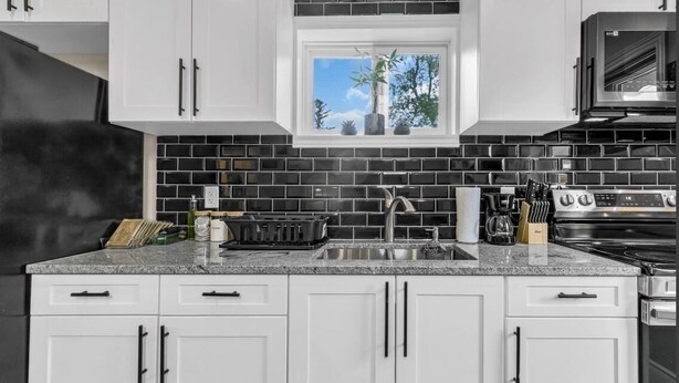 A kitchen with white cabinets, a black tiled backsplash, a window with a view of trees, and a stainless steel stove.