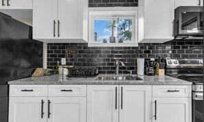Une cuisine avec des meubles blancs, un dosseret en carreaux noirs, une fenêtre donnant sur la verdure et une cuisinière en acier inoxydable.