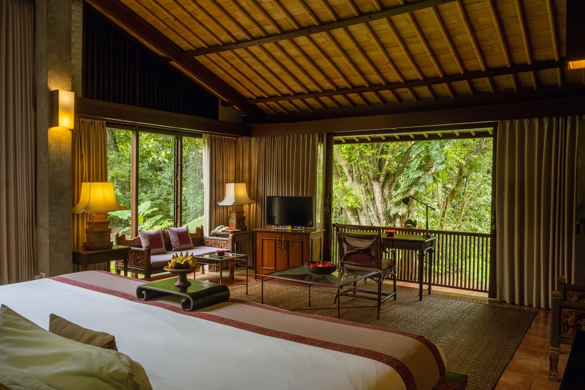 A spacious bedroom with a large bed, a desk with a television, and a balcony with a view of the surrounding forest.