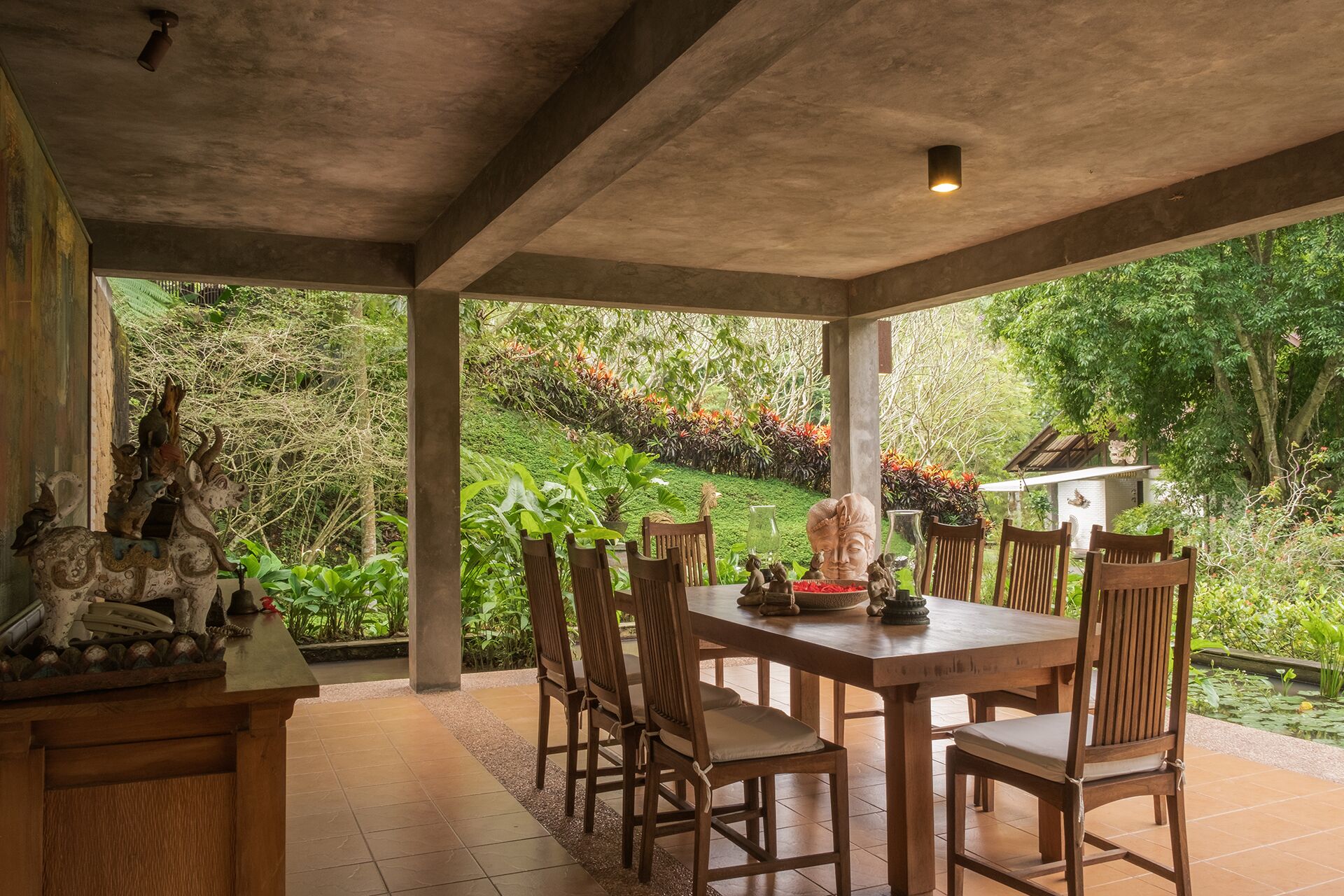 A covered outdoor dining area with a table and chairs, surrounded by lush greenery and a small pond.