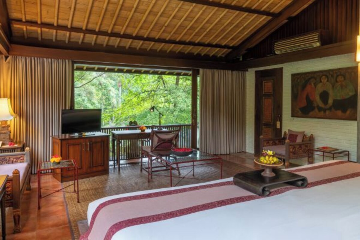 A hotel room with a bed, a TV, a desk with a chair, a dining table with chairs, and a large window with curtains.