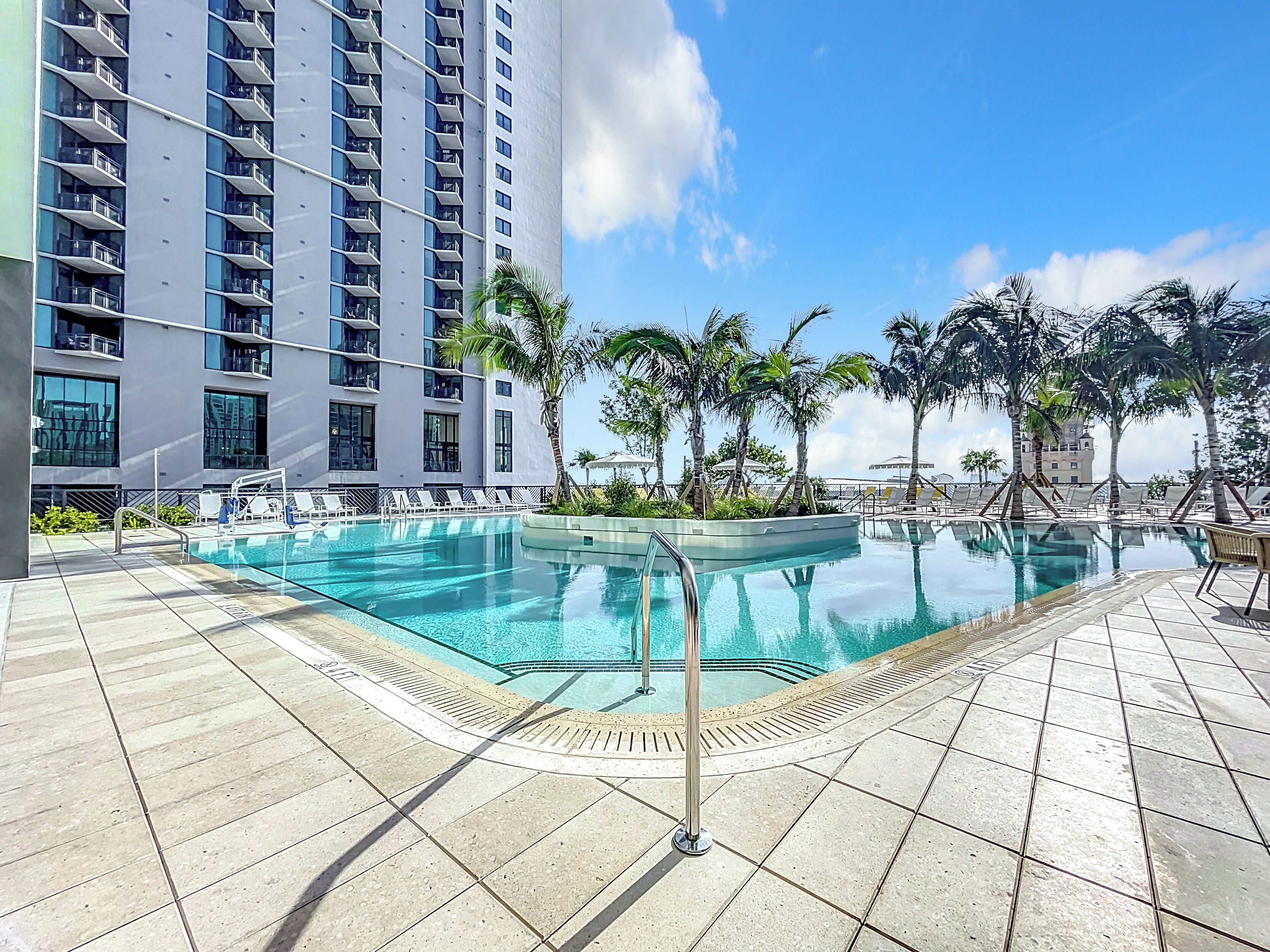 A swimming pool with lounge chairs and palm trees, surrounded by a high-rise building.