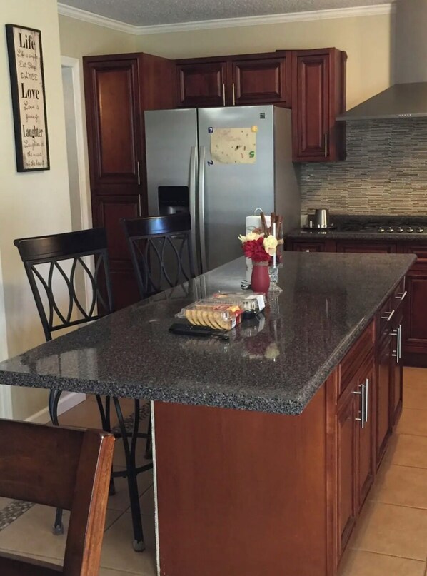 A kitchen with dark wooden cabinets, a stainless steel refrigerator, a granite countertop, and a dining area with chairs.