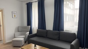 A modern living room with a grey sofa, a grey armchair, and a small ottoman. There is a wooden coffee table with a plant. Blue curtains cover the windows, and a framed picture hangs on the wall.