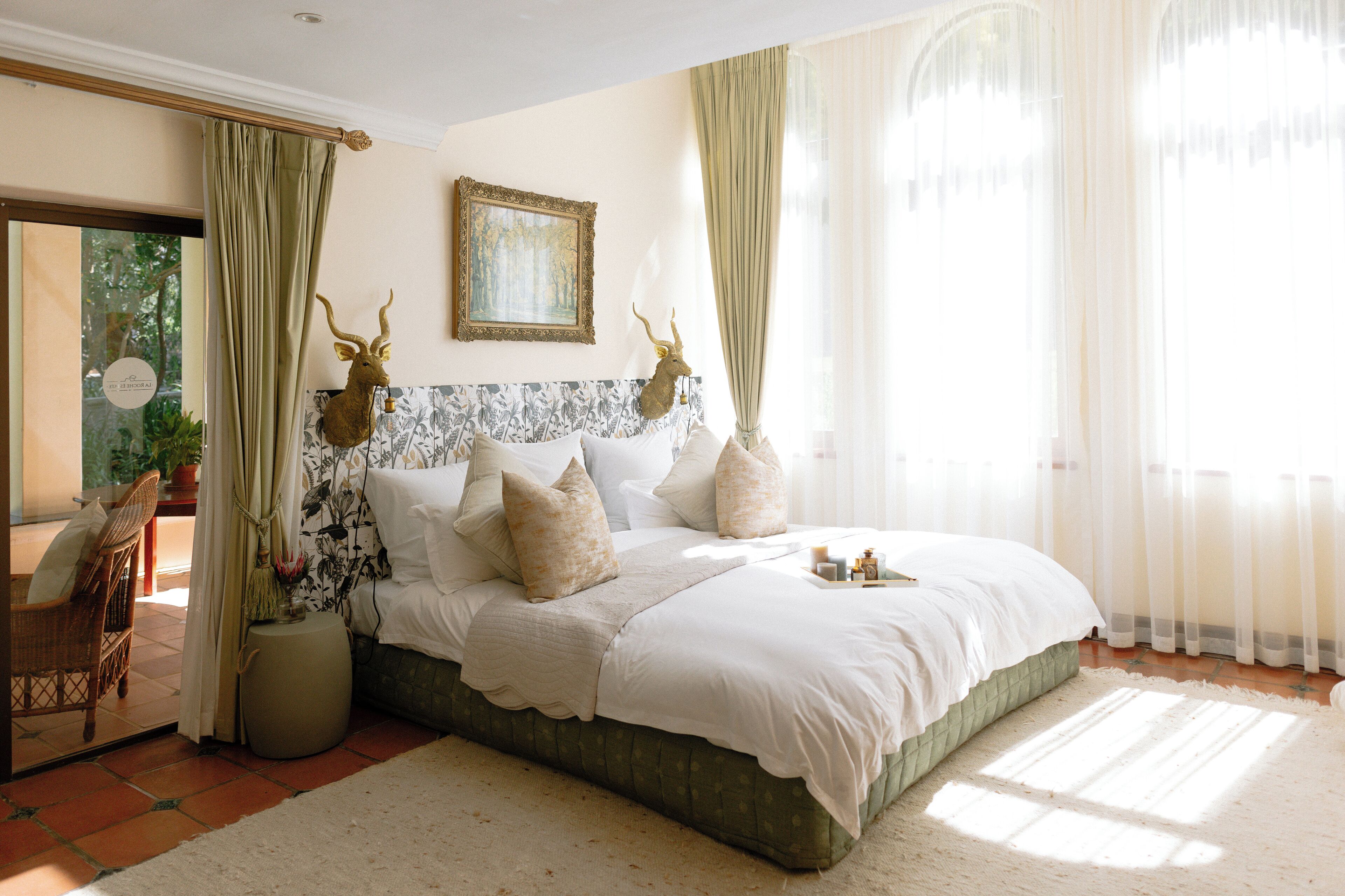 Een slaapkamer met een groot bed, een hoofdeinde met gewei, een lijst met een schilderij en uitzicht op groen buiten.