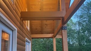 A wooden deck with a covered seating area, a hanging light fixture, and a view of the surrounding forest. - Luxury 3-bed hemlock log cabin Cave Run Lake area, with WiFi, gameroom, hot tub (West Liberty)