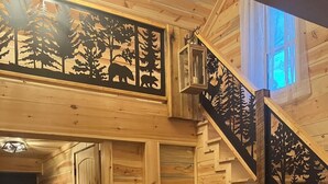 A wooden interior with a staircase featuring carved tree designs, a bear sculpture, and a view of the outdoors. - Luxury 3-bed hemlock log cabin Cave Run Lake area, with WiFi, gameroom, hot tub (West Liberty)