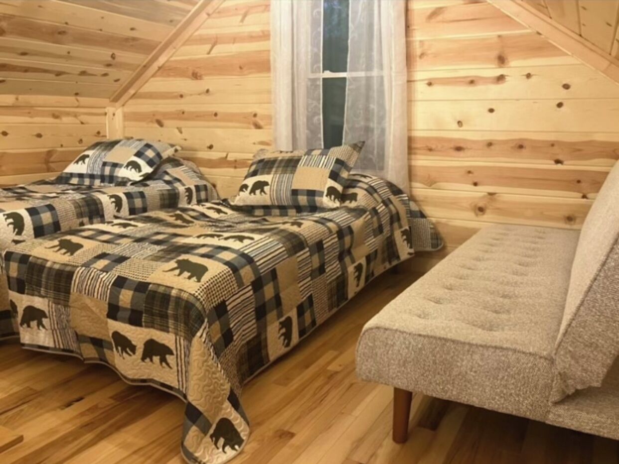 A cozy cabin with two beds covered in a bear-patterned blanket, a bench, and a window with curtains.