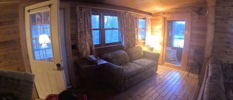 A cozy cabin interior with a wooden floor, a sofa, a door, and a window with curtains.