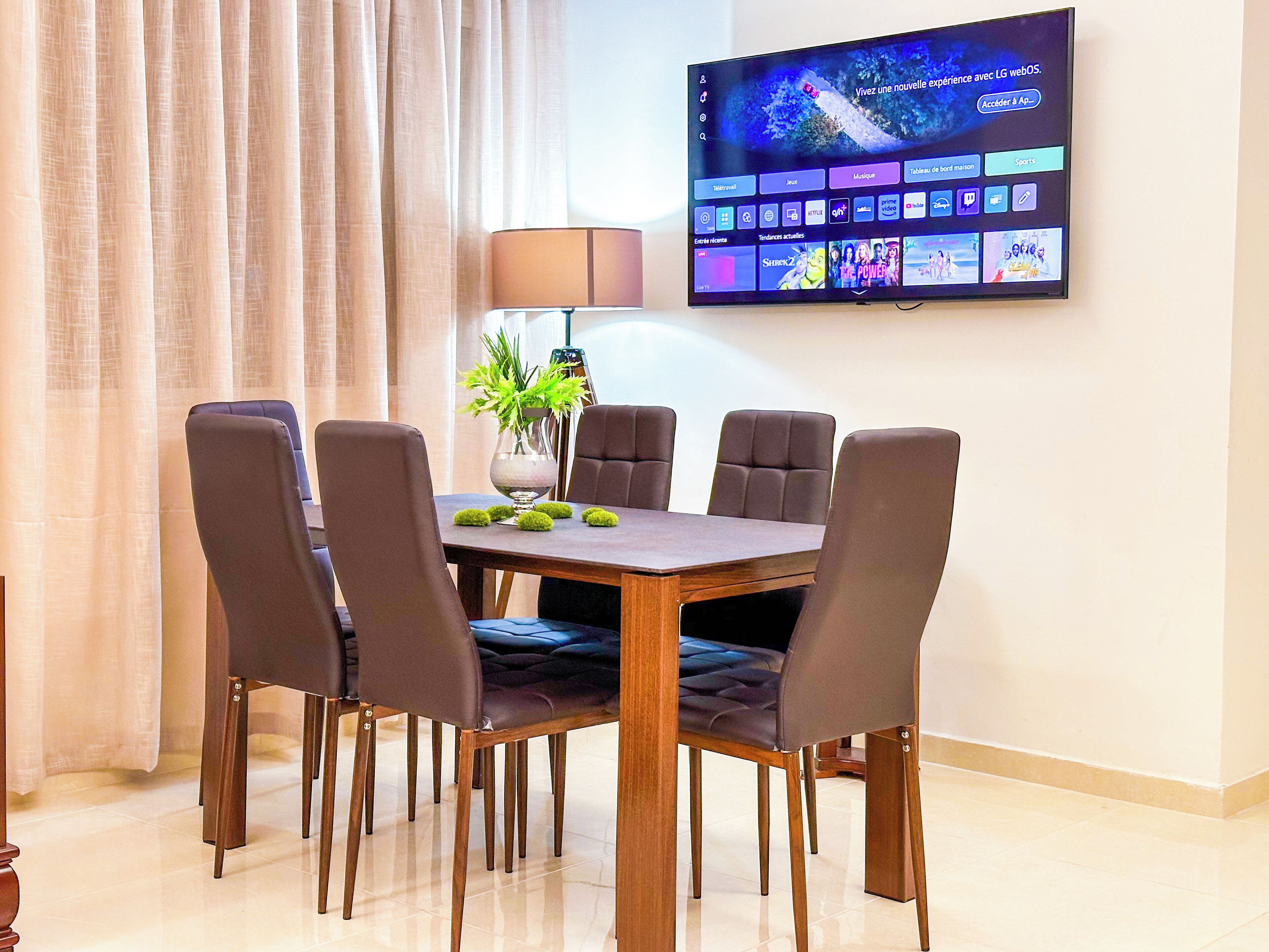 A dining area with a wooden table and six chairs, a vase with green plants, and a flat-screen TV mounted on the wall displaying a streaming service interface.