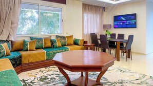 A modern living room with a sofa, a dining table, chairs, a TV, and a chandelier. - Luxoria home - 2 min to corniche (Tangier)