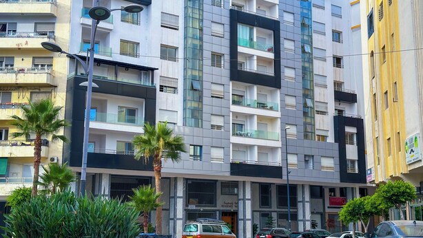 A multi-story residential building with balconies, a parking area, and a small garden with pink flowers. - Luxoria home - 2 min to corniche (Tangier)