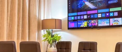 A hotel room with a flat-screen TV mounted on the wall displaying a streaming service interface. There is a dining table with four chairs and a potted plant on it.