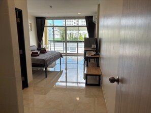 A hotel room with a bed, a desk, a chair, and a large window with a view of buildings and trees. - new sorot  house  (Pattaya)