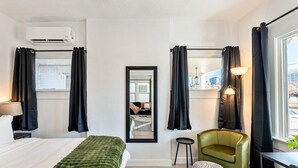 Una habitación de hotel moderna con una cama, un sillón verde, un taburete pequeño, un espejo y un equipo de aire acondicionado.