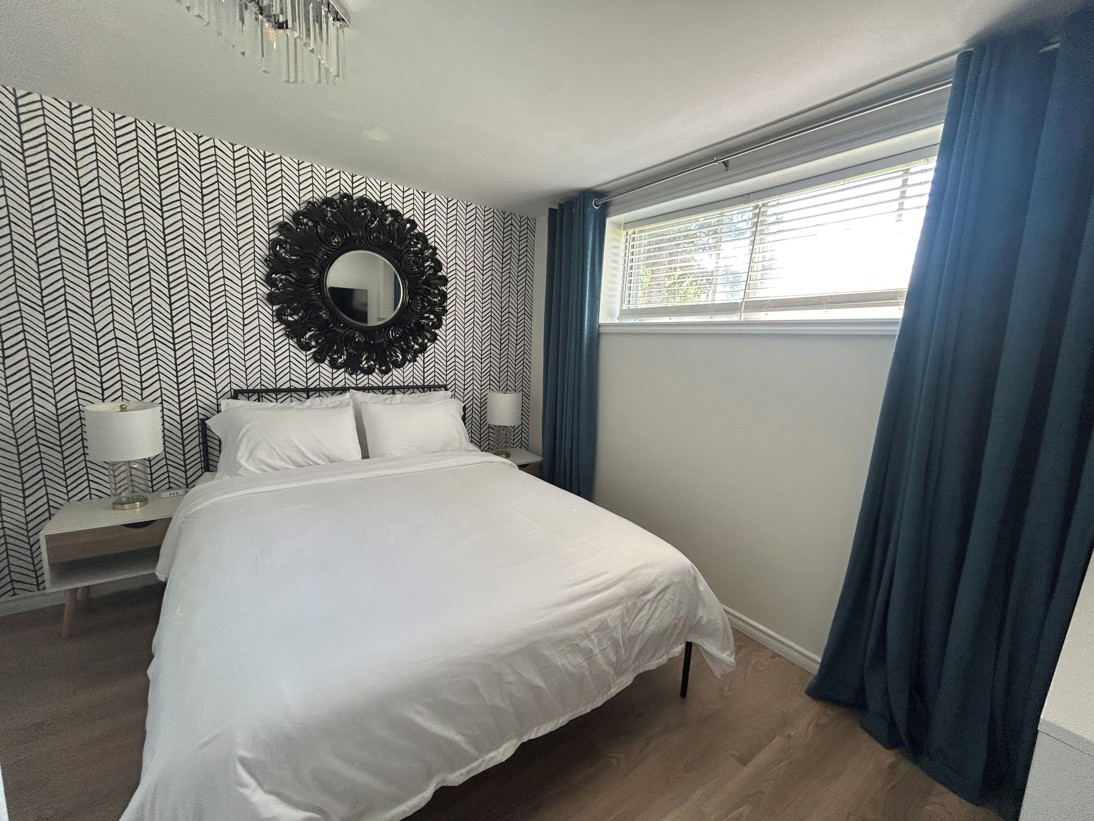 A bedroom with a bed, a round mirror, two bedside tables with lamps, a window with curtains, and a patterned wallpaper.