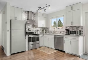 A modern kitchen with white cabinets, stainless steel appliances, a patterned backsplash, and a window providing a view of trees. - Large Groups, Sleeps 16, AC, King Bed, Fire Pit, large patio. (Calgary)