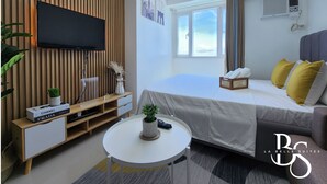 Una habitación de hotel moderna con una televisión de pantalla plana, una cama con almohadas amarillas y grises, una mesita redonda con una planta y un panel de pared de madera.