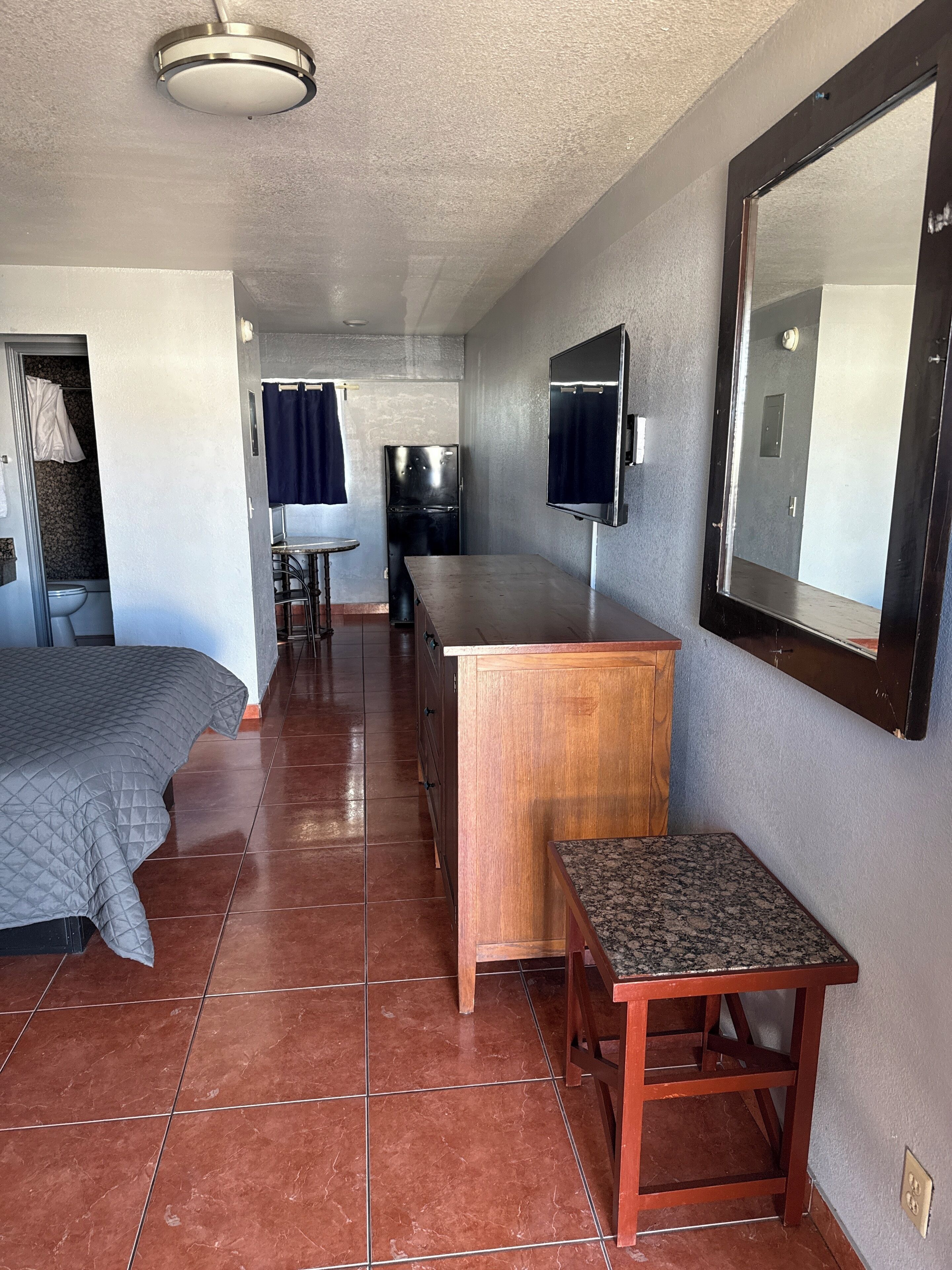 A compact hotel room with a bed, a small wooden desk, a chair, a TV mounted on the wall, and a bathroom visible through an open door.