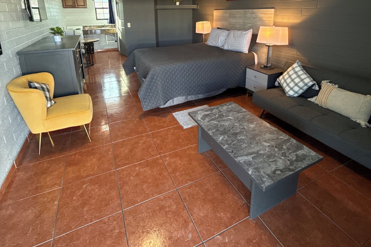 Une pièce compacte et moderne, comprenant un lit, un canapé, un petit fauteuil et une table basse. La pièce est dotée d’un sol carrelé et d’un luminaire au plafond.