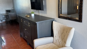 Uma sala de estar moderna com uma televisão de tela plana na parede, um armário escuro, uma poltrona branca com capitonê e uma área de jantar com mesa e cadeiras.