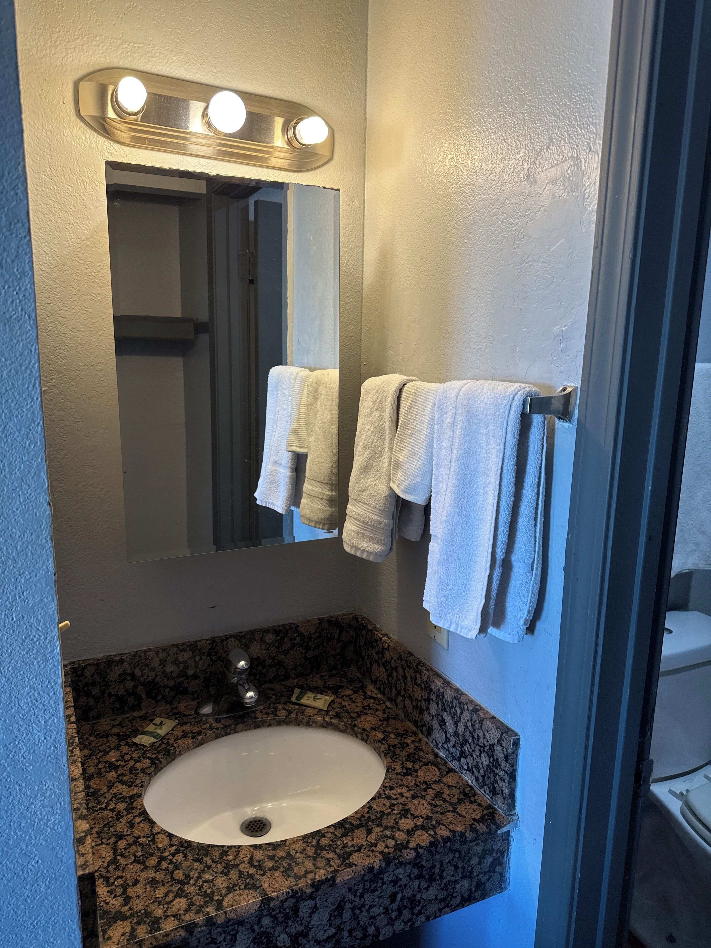 A bathroom with a granite countertop sink, a mirror with lights, and two towels hanging on a rack.