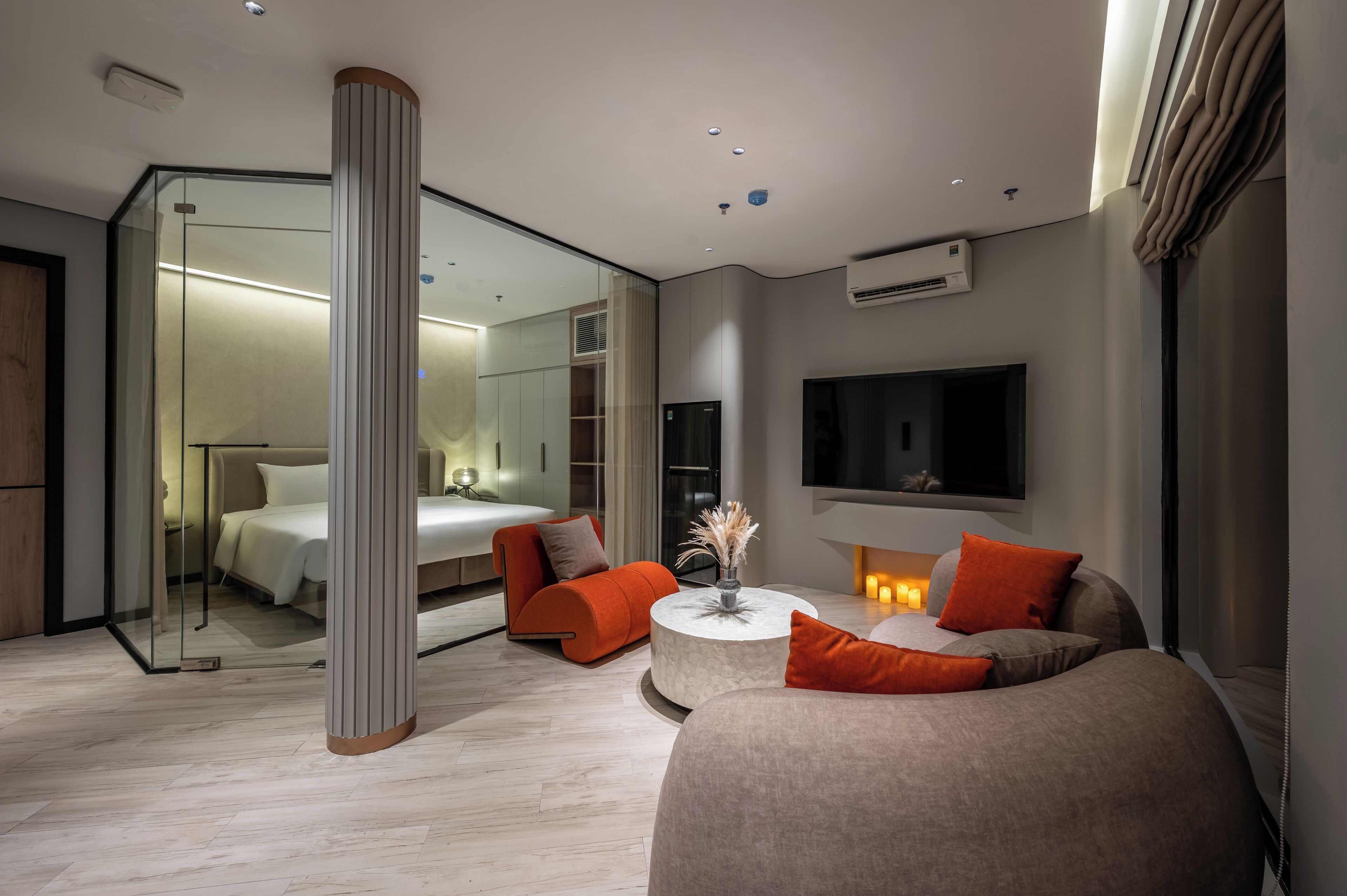 A modern hotel room with a glass partition, a round coffee table, a sofa, and a TV mounted on the wall.