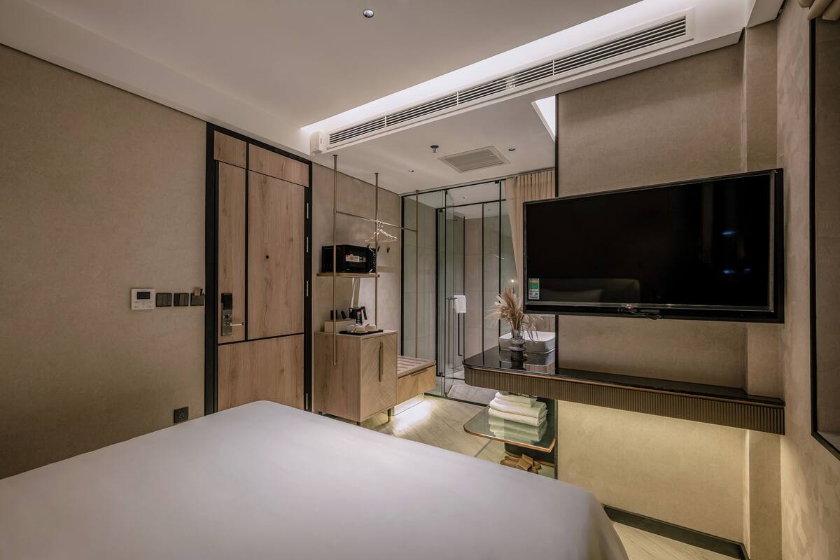 A modern hotel room with a large bed, a TV mounted on the wall, a bedside table with a lamp, and a small seating area with a chair and a table.