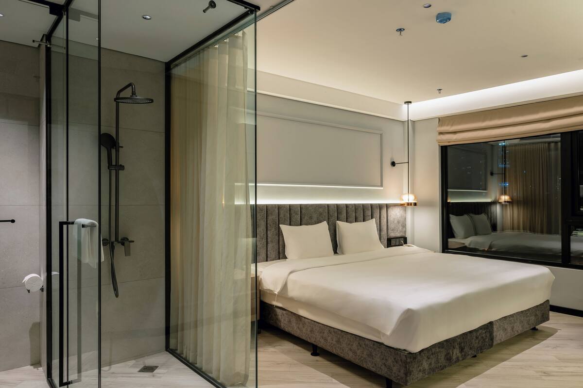 A modern hotel room with a glass-enclosed shower, a large bed with a headboard, and a view of the city through a large window.