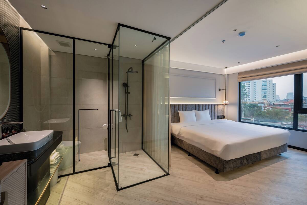 A modern hotel room with a glass-enclosed shower, a large bed, a vanity with a sink, and a view of the city through a large window.