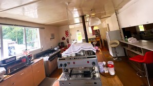 A compact kitchen with a coffee machine, a stove, and a sink. There is a window providing natural light, and a dining area with a red chair and a table. - Taringatura Backpackers (Dipton West)