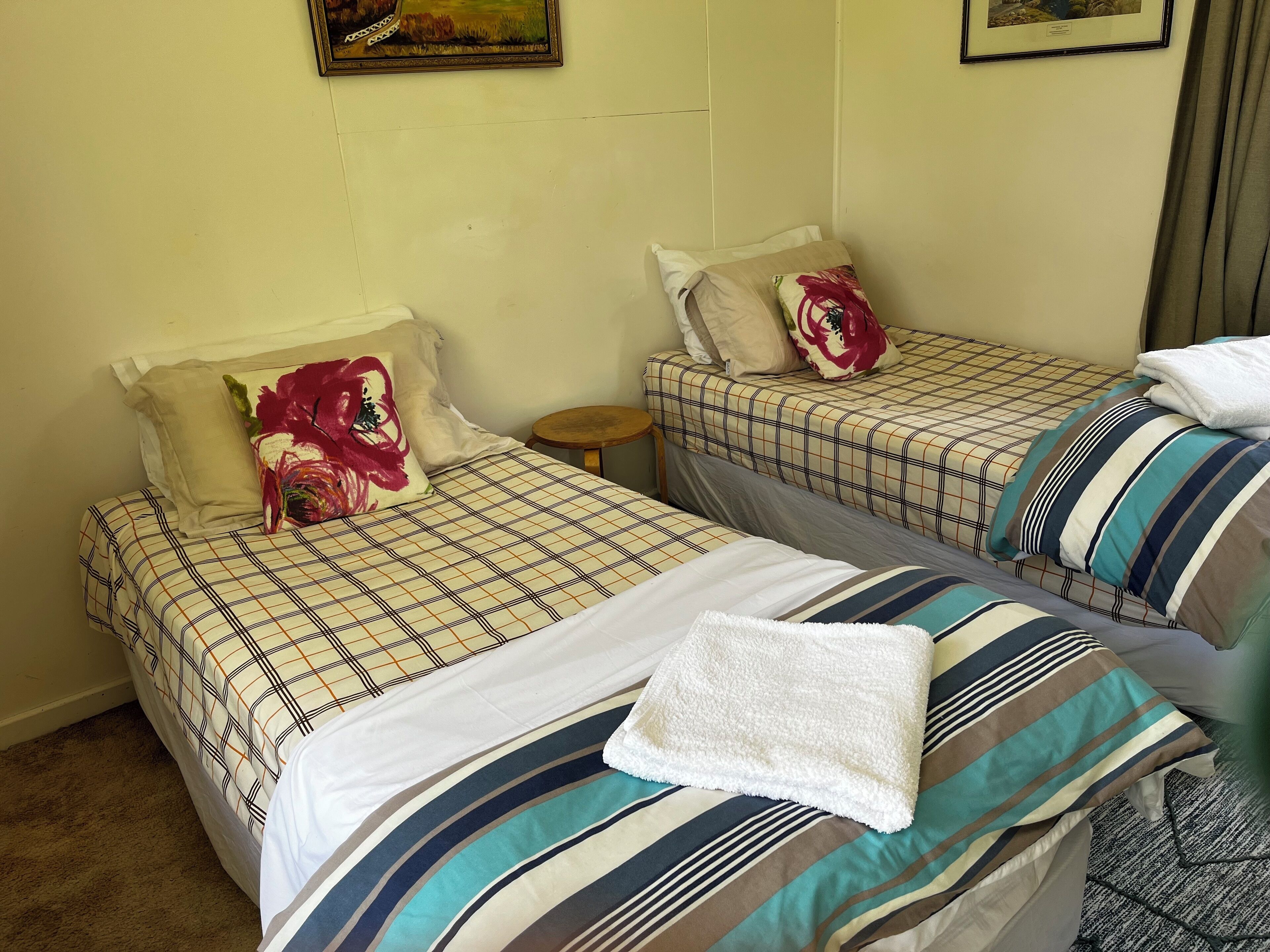 A room with two beds, each with a plaid bedspread and a pillow with a floral design. There is a small wooden stool beside one bed, and a framed picture on the wall.