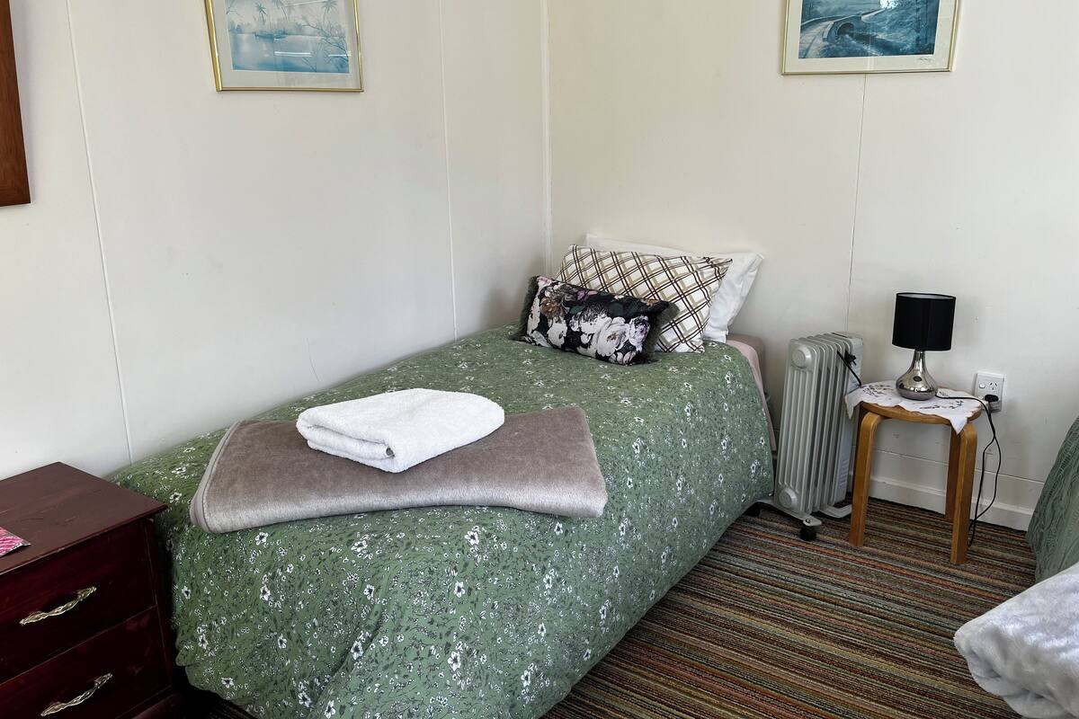 Ein ordentlich bezogenes Bett mit einem grün-floralen Überwurf, einem weißen Handtuch und einer braunen Decke. Ein hölzerner Nachttisch mit einer Lampe und einem Heizkörper stehen neben dem Bett. Zwei gerahmte Bilder hängen an der Wand über dem Bett.