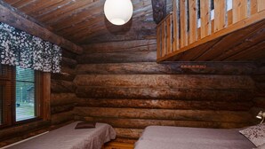 Un dormitorio tipo cabaña de troncos con dos camas, una lámpara colgante, vigas de madera y una ventana con persianas.
