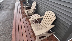 A wooden deck with three Adirondack chairs, a metal table, and a house with grey siding.