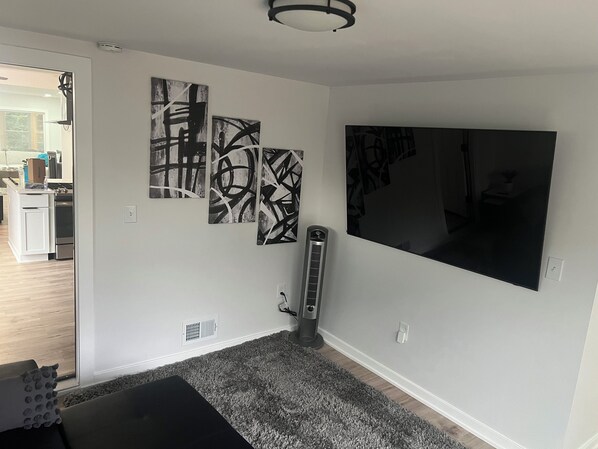 A living room with a large flat-screen TV, abstract wall art, a standing heater, and a grey carpet.