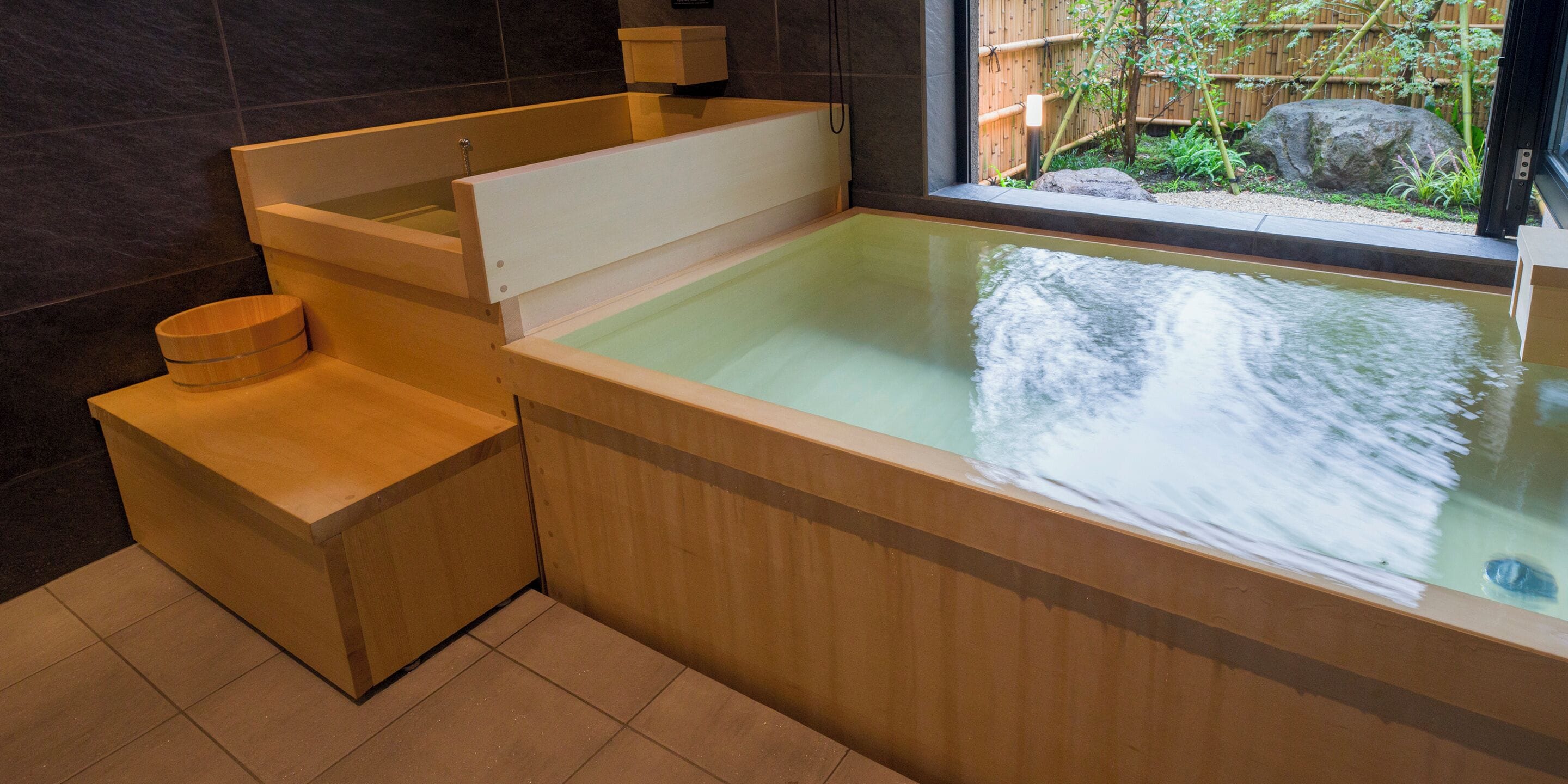A modern bathroom with a wooden bathtub, a wooden bench, and a window providing a view of a bamboo fence and rocks.