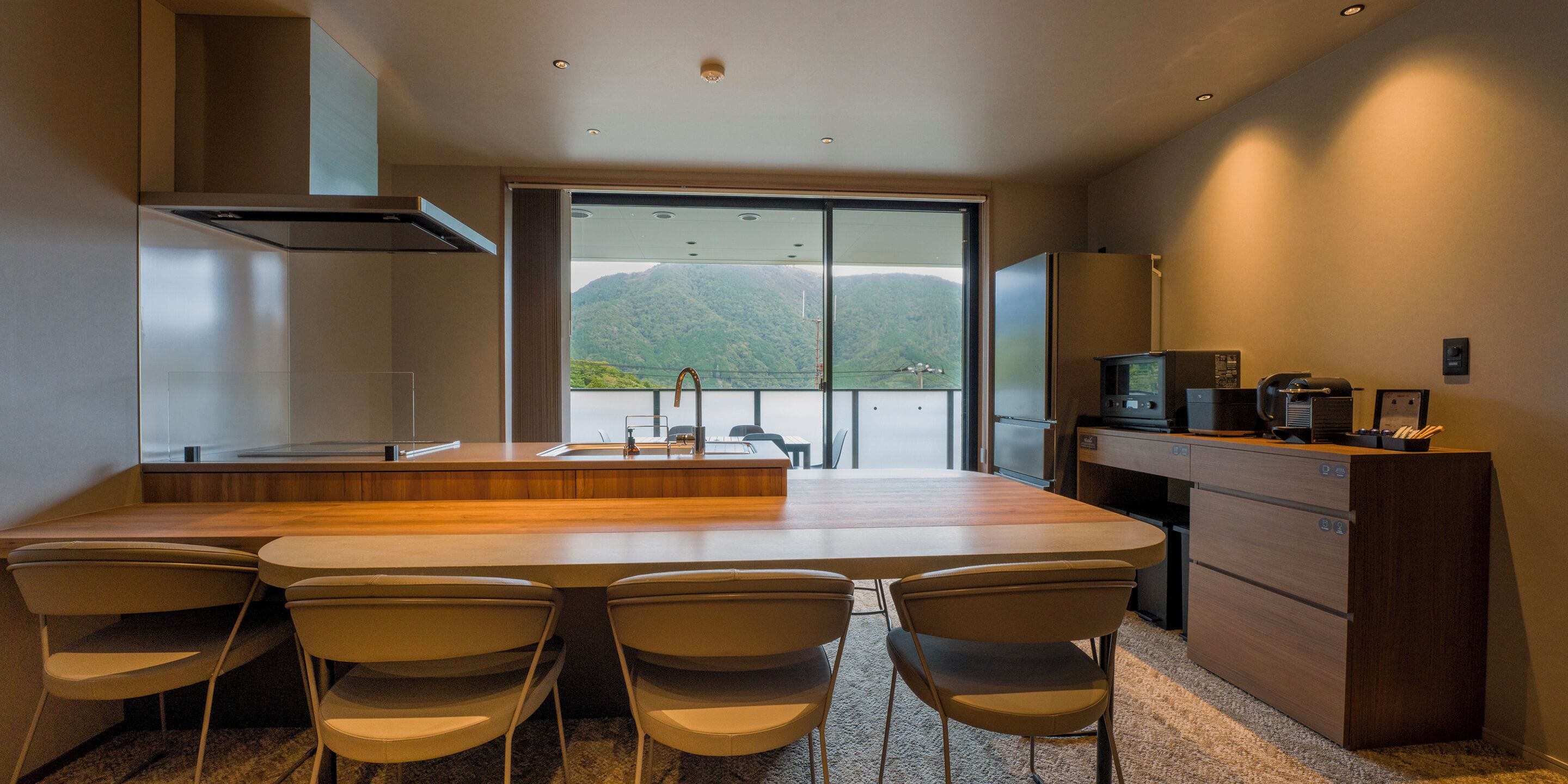 Una cucina moderna con una grande isola, quattro sedie, forno a incasso e vista sulle montagne attraverso un'ampia finestra.