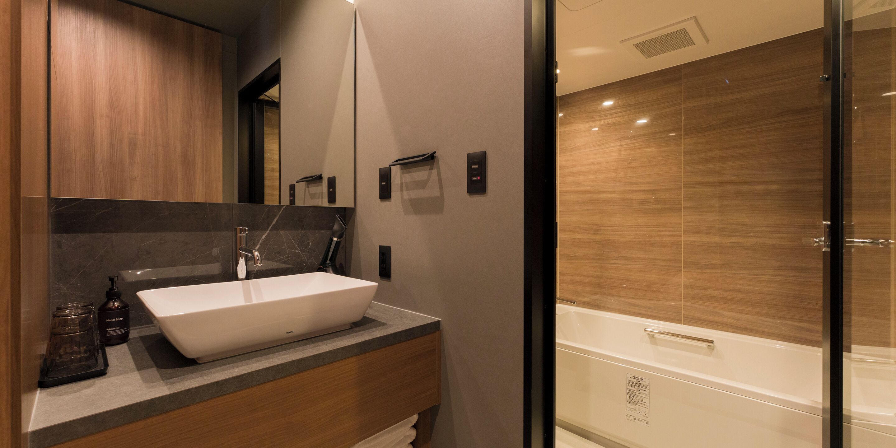 A modern bathroom with a large white sink, dark marble countertop, wooden cabinets, and a glass-enclosed shower.