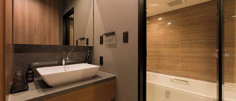 A modern bathroom with a large white sink, dark marble countertop, wooden cabinets, and a glass-enclosed shower.