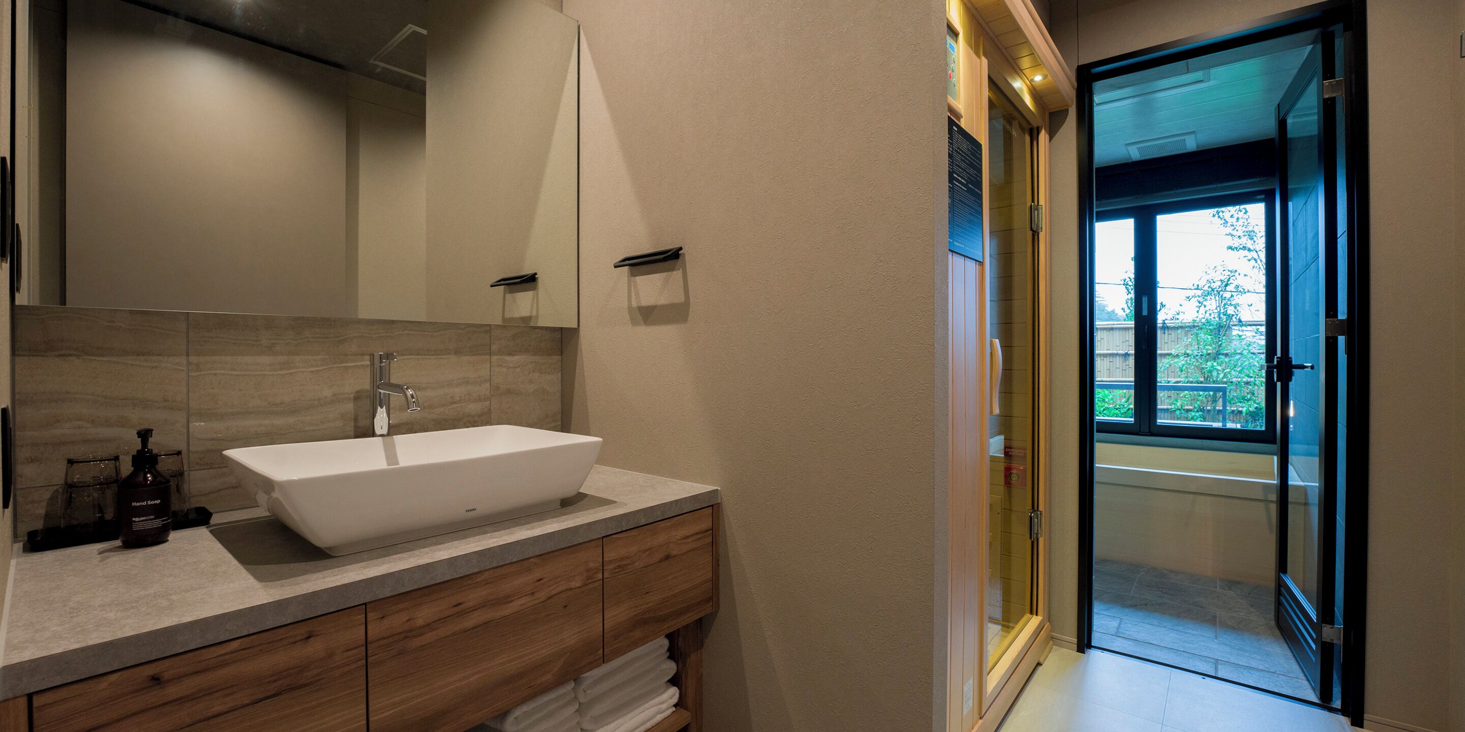 Een moderne badkamer met een ruime wastafel, een houten wastafelmeubel en een glazen deur naar een buitenruimte.