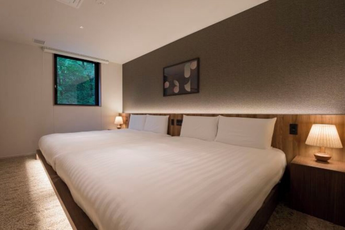 Een moderne hotelkamer met een groot bed, een nachtkastje met lamp, een raam met uitzicht op bomen en een kunstwerk aan de muur.